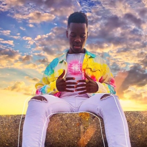 Tashinga Bepete sat on a wall against a background of clouds and blue sky at sunset. He is wearing a bright multicoloured jacket, white top and white jeans.